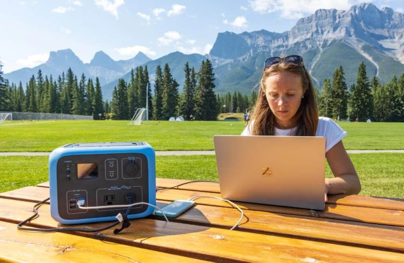 A woman using a portable solar generator to work on a laptop in a mountainous forest setting